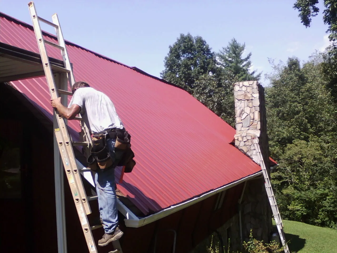 Roofer on a ladder beside a red metal roof and stone chimney — Soffit & Fascia Repair by Roofing Brothers in Dauphin